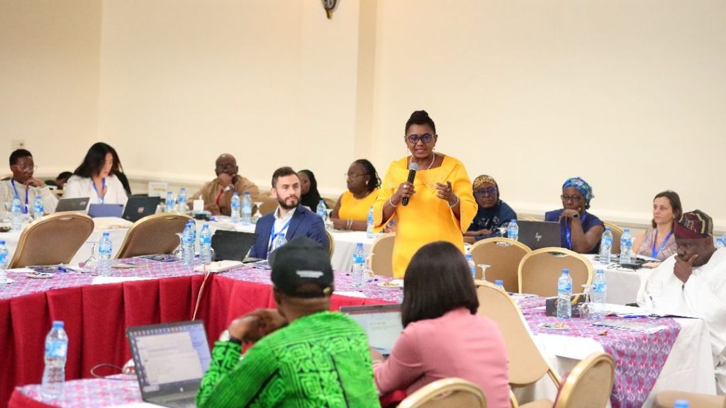 A woman in a yellow dress stands and speaks into a microphone at a roundtable event